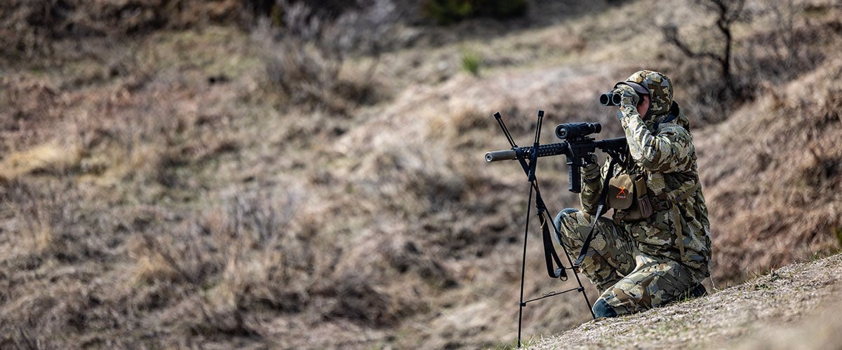 hunter looking through binoculars while kneeling on the side of a hill